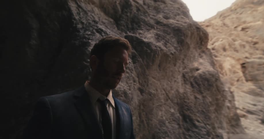 A man in a suit looks around while standing near a cave in a canyon