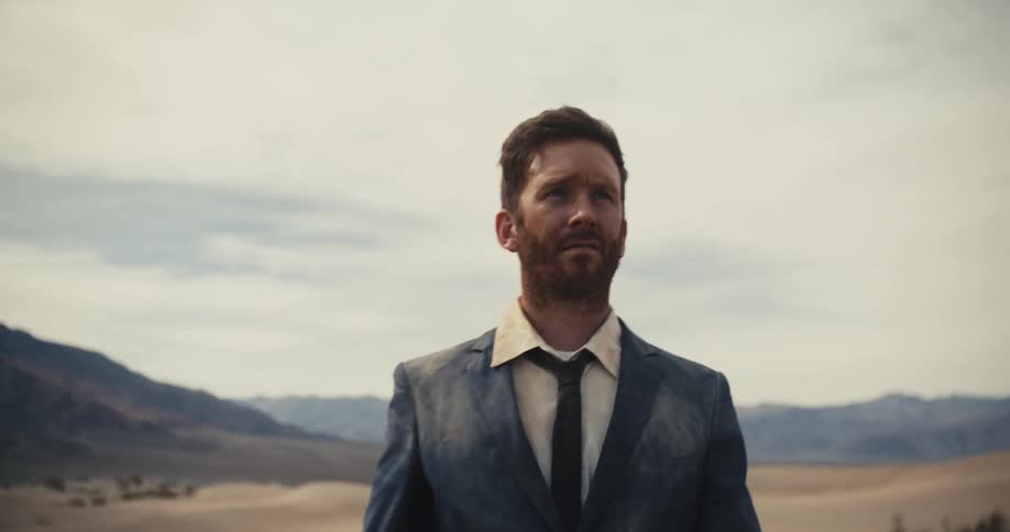 The sun is shining on the face of a man in a dirty suit walking in the desert looking tired and stumbling