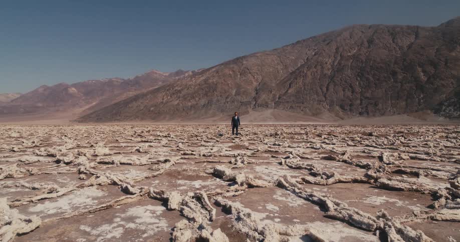 A man wearing a suit is walking in the desert and there are mountains in the distance under a clear sky