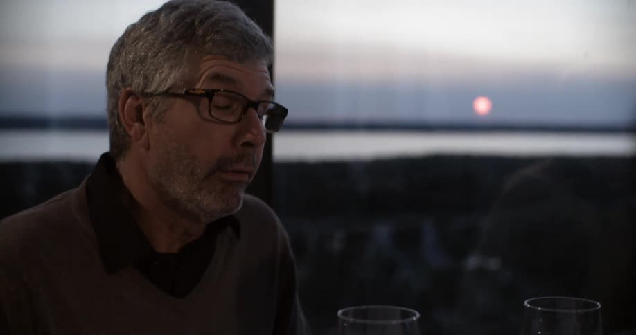 An older man wearing glasses has a chat over dinner near a window at dusk