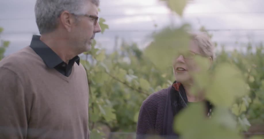 An older man and woman walking through grape vines and talking