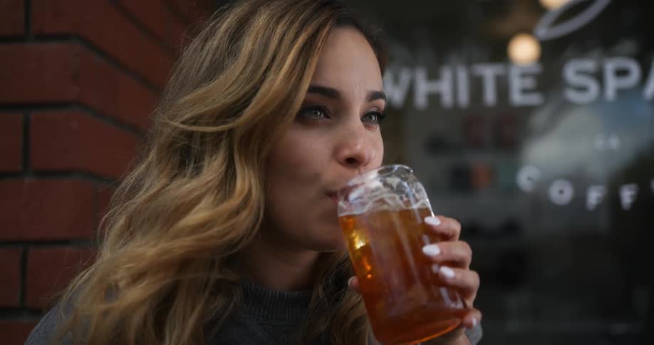 Woman with brown wavy hair and green eyes sits at table in front of cafe drinking iced coffee