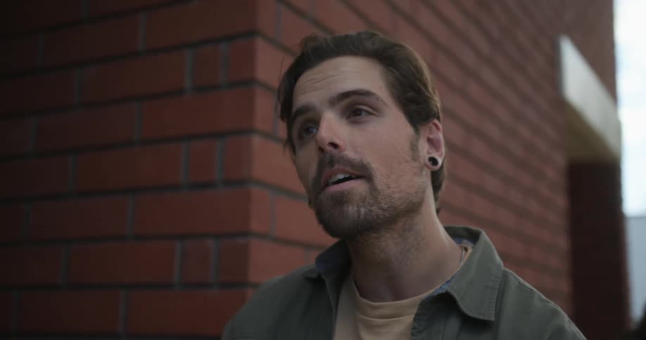 Man with stubble beard green shirt and ear tunnel plugs drinks coffee from cup in front of cafe talking