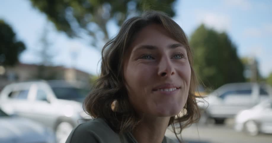 Woman sitting at outdoor cafe smiles and picks up coffee as traffic passes by in background