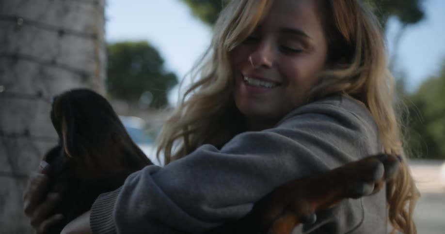 A woman is outside hugging her dog as a truck passes by on the street in the distance