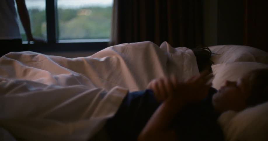 Two people lay in a bed while someone stands near the bed looking out of a window