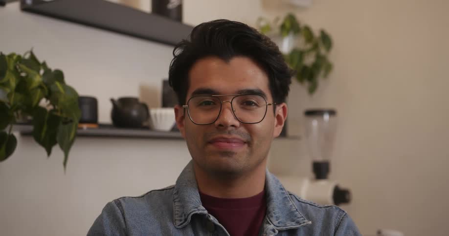 Barista wearing glasses and denim jacket smiles at camera in cafe