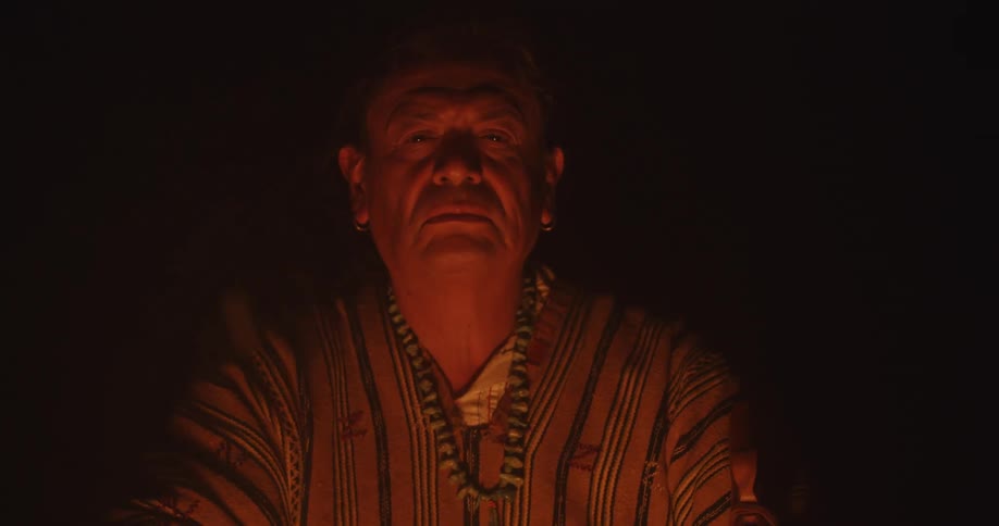 A man is sitting outside relaxing as light from his campfire shines on his face as sparks and smoke rise in the darkness