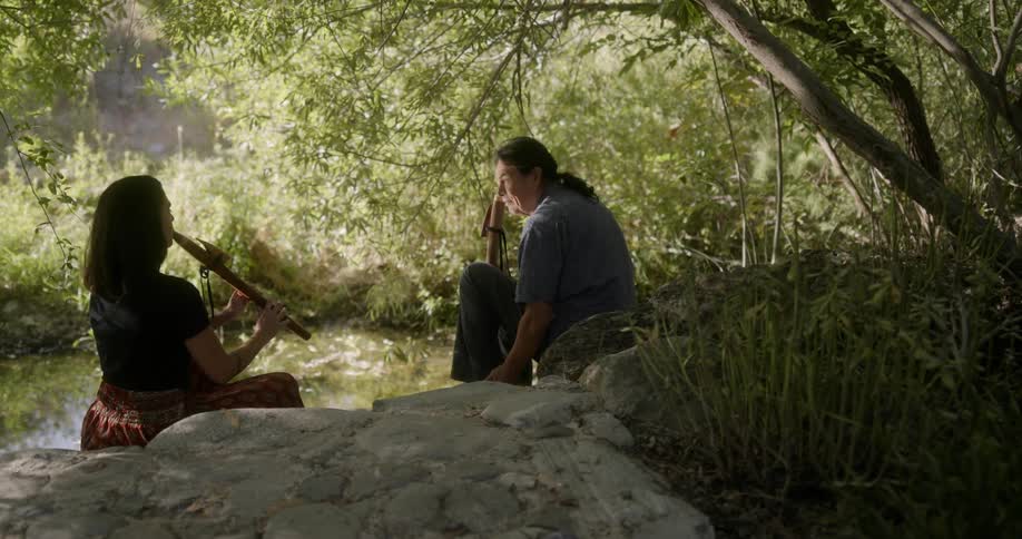 A man holding a woodwind instrument looks at a woman playing the same instrument as they sit together at the side of a pond during the day