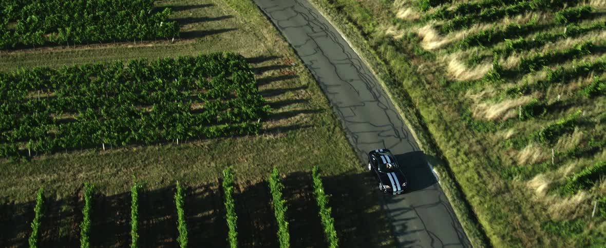 A sports car travels down a narrow road through a farmland during the day