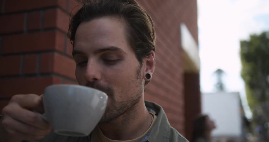 Man wearing green shirt and ear tunnel plugs drinks a cup of coffee in front of cafe talking to person
