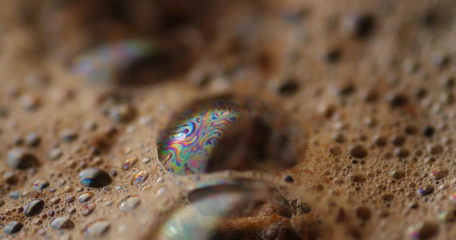 Iridescent colors move across surface of large bubble in foam of coffee