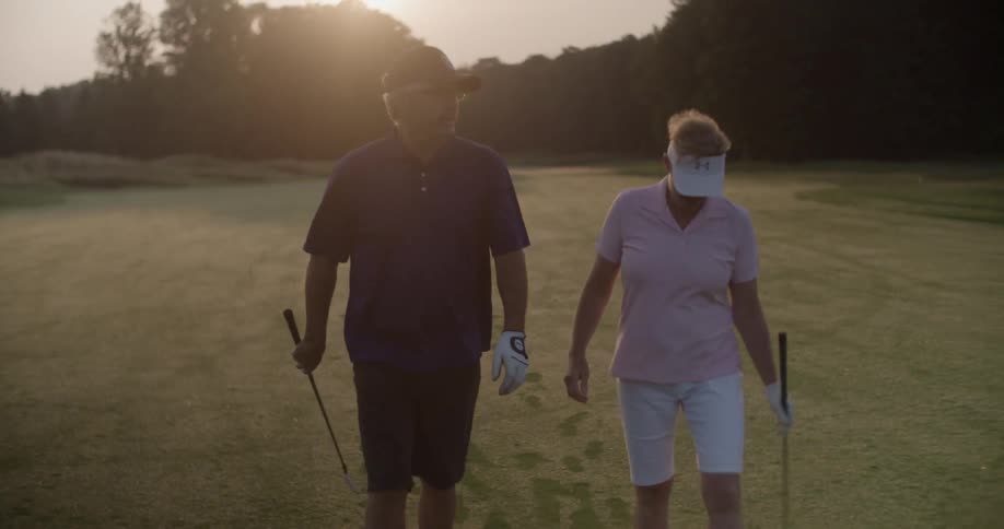 A man and woman walk on a golf course at dusk while carrying golf clubs