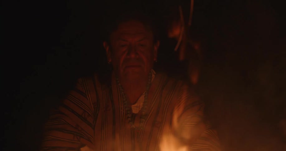 A man is sitting near a campfire watching the sparks and flames rise among the smoke in the darkness