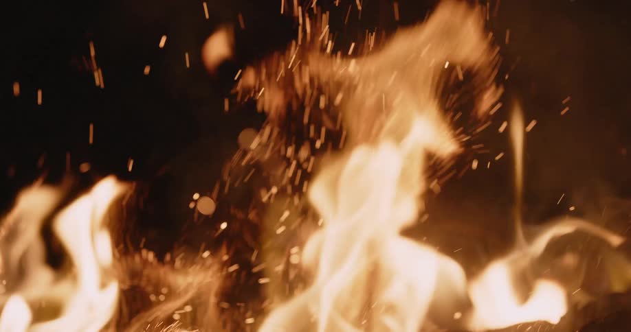 The flames of a campfire waver as they move and sparks rise in the darkness