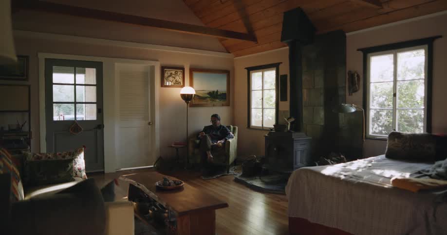 Man with long hair and glasses sits in armchair reading a book in room
