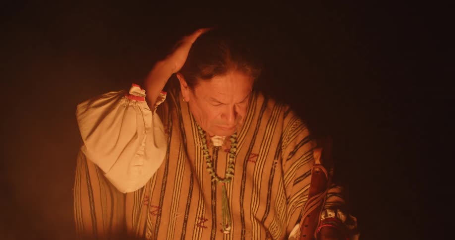 Fire light shines on a native american man as he is sitting in front of a campfire performing a fire ritual in the darkness