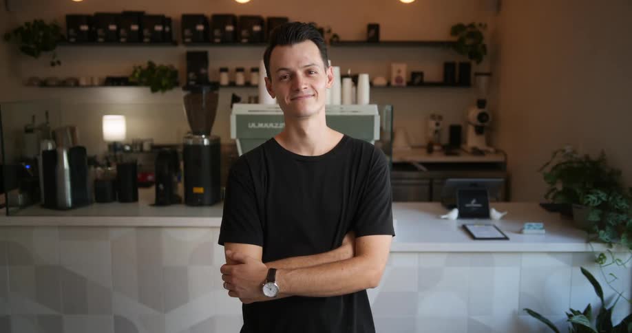 A young man is standing inside a cafe as he folds his arms and smiles