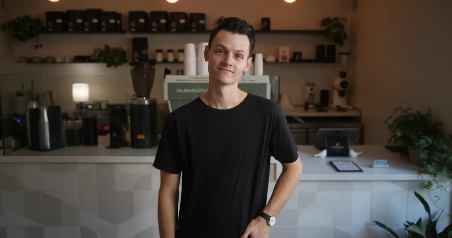 Barista with black t shirt and wristwatch looks at camera in front of cafe bar
