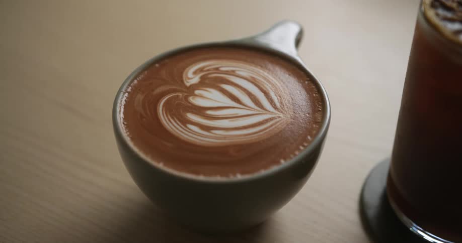 Latte decorated with coffee art sits on table next to iced coffee in a glass