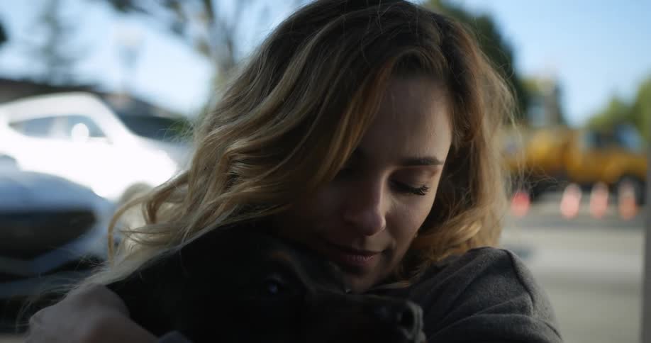 Woman with brown hair and gray sweater kisses and pets black dog in the street