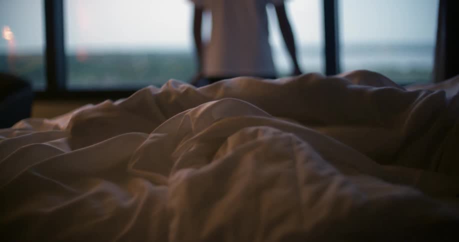 A man looks out the window as another person is in bed