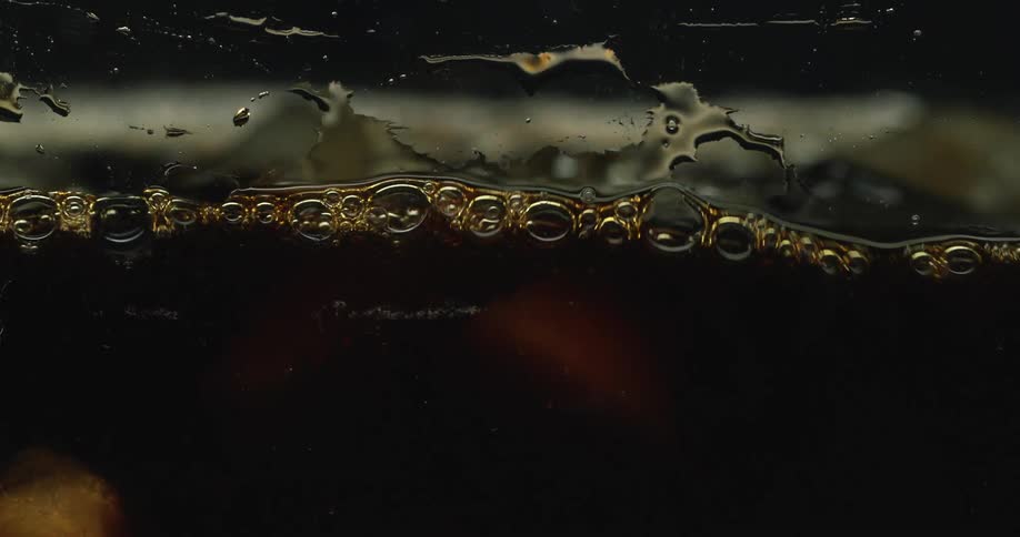 Ice cubes are dropped into dark beverage creating a splash and bubbles