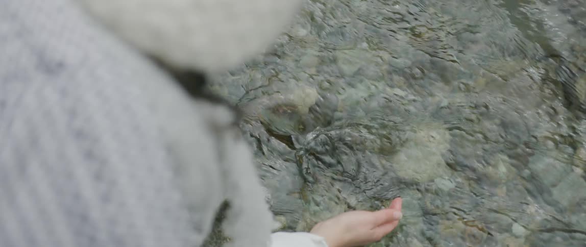 A woman is standing over a clear running stream as she reaches into the water and pick up stones and lets water rush into the palm of her hand