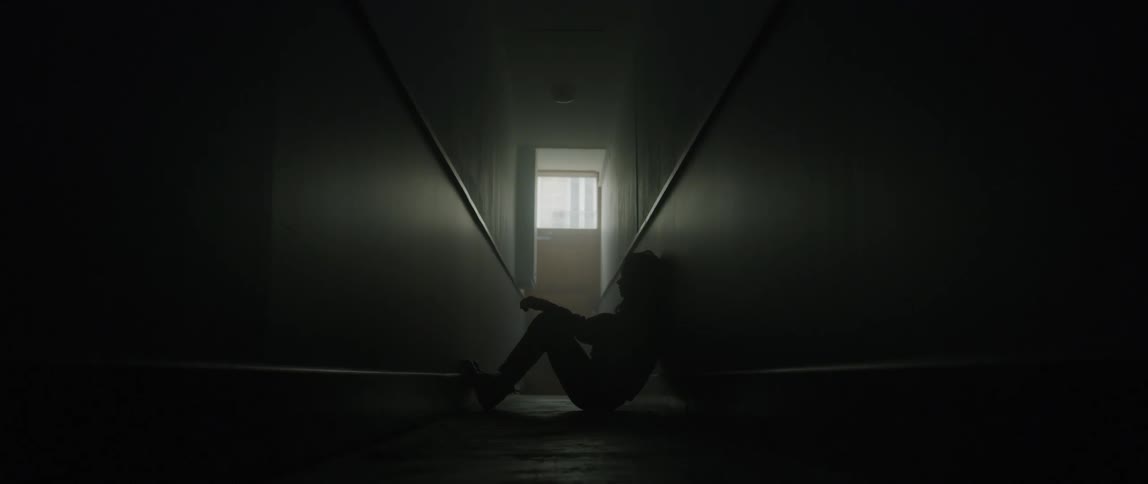 A sad female standing in a hallwayslides down a wall and sits