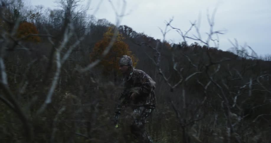 A hunter carrying a crossbow climbs a steep hill covered in dense vegetation on a cloudy day