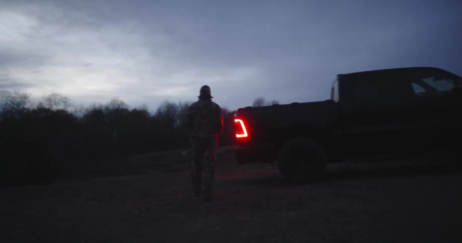 A hunter stores a crossbow in the back of a truck parked in a grassland on a cloudy evening