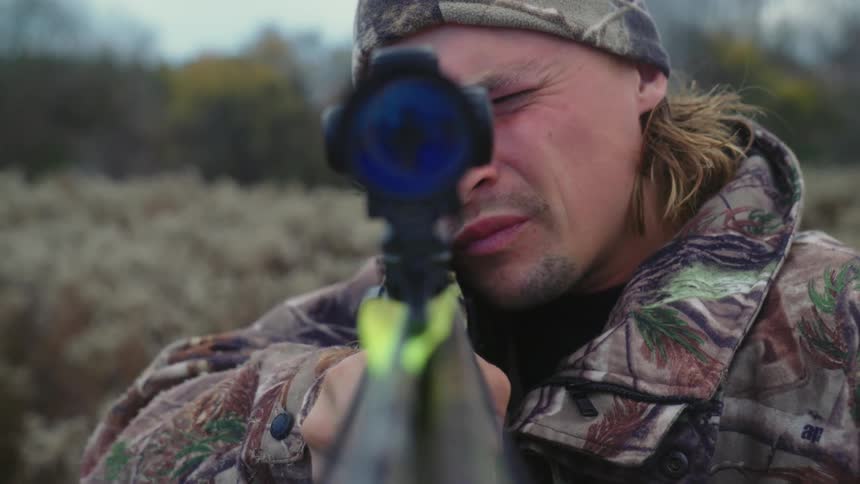 A man is standing outside holding a crossbow as he bends down to look through the sight