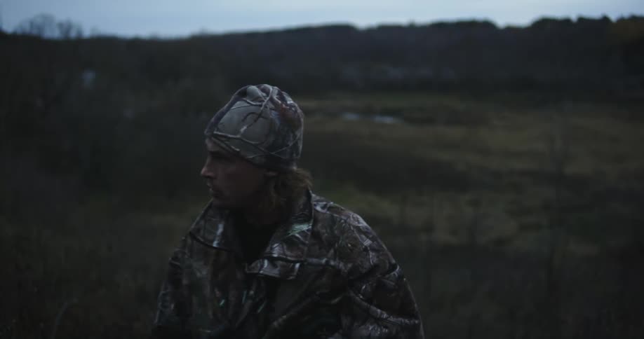 A hunter carrying a crossbow climbs a hill and stops before the camera while looking out of frame