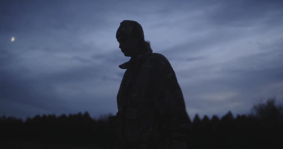 A teenage boy wearing a coat and hat carrying something as he walks near woods as the moon shines through the clouds in the sky