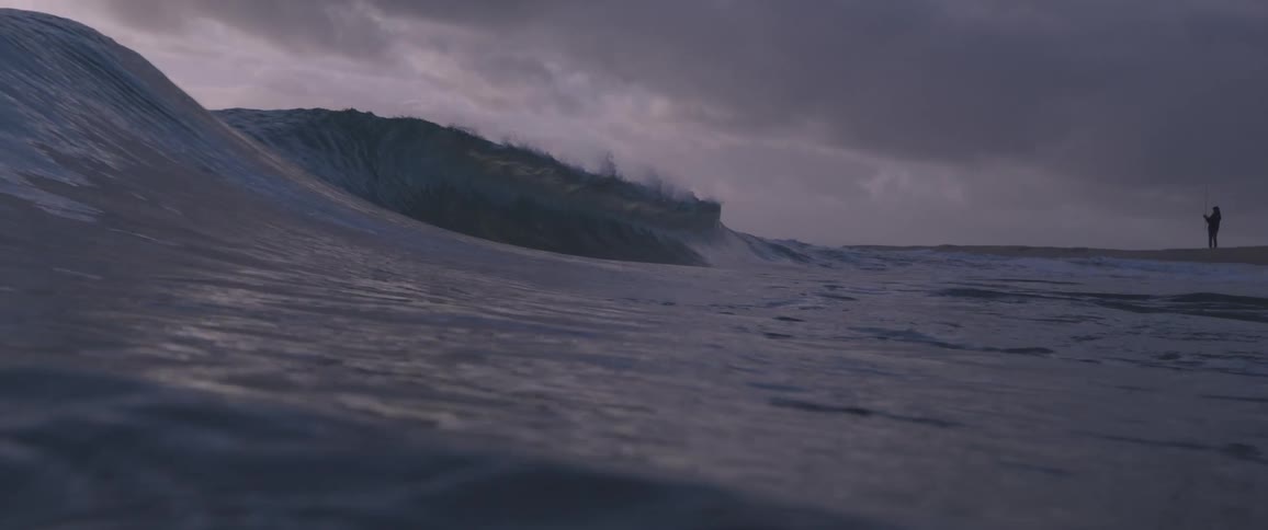 Large wave rolls across ocean as people stand on beach in the background