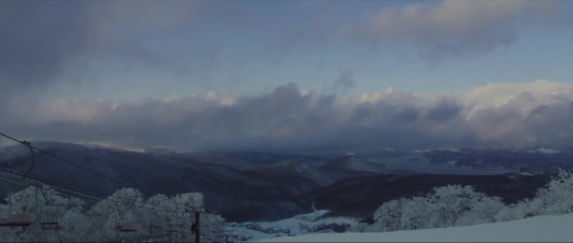 Lone person rides ski lift overlooking snowy mountain range on a cloudy day