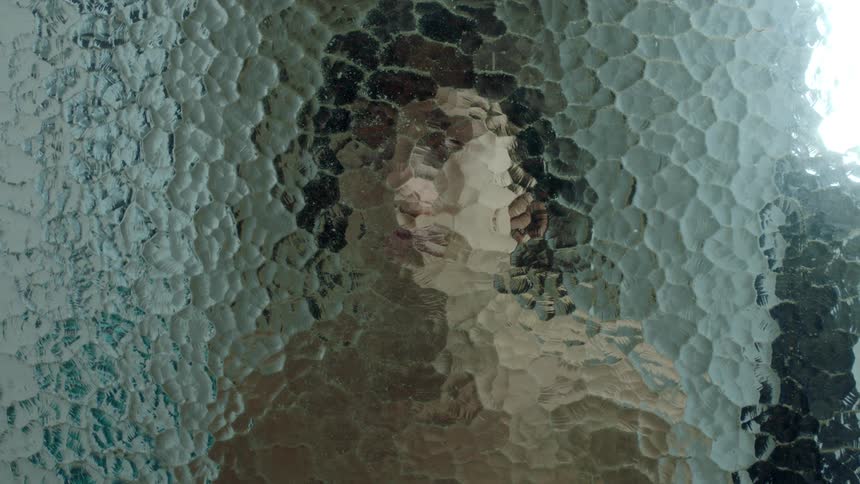 A woman standing behind glass lifts her hand to touch the glass