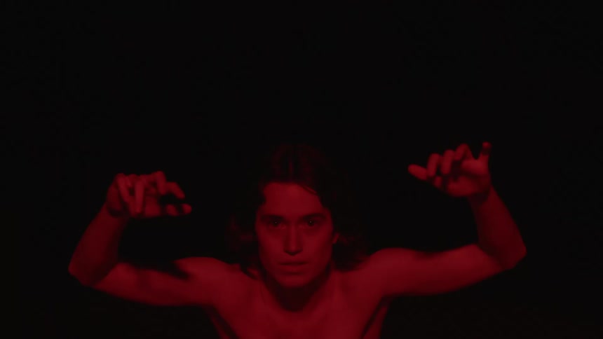 Red light shines on a bare chested man with his arms in the air in the darkness