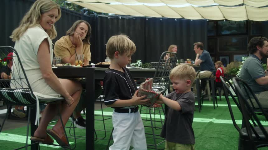 Two littles blonde boys playing with a baseball glove while a woman and man sit at the table next to them watching and smiling