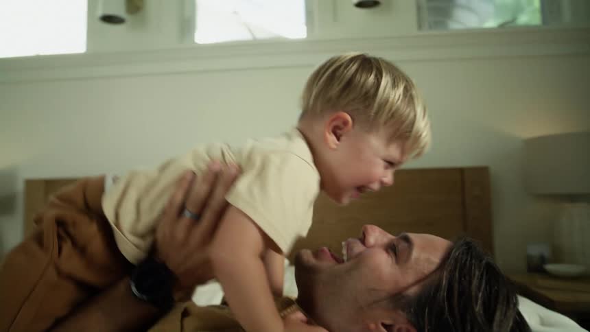 A man lies on his bed at home and his young son steps on him and he picks him up and plays when his other son comes and hits him with a pillow as they all laugh