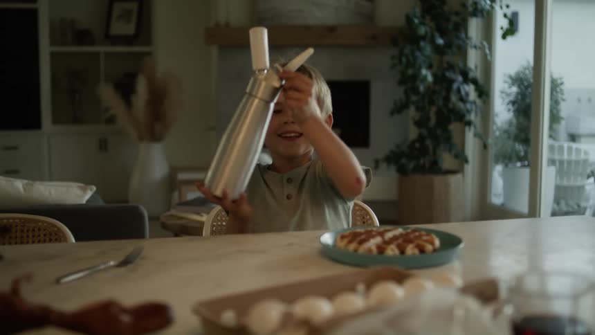 A dad making waffles at home puts a waffle on a plate and hands it to his young son who squirts whipped cream on top