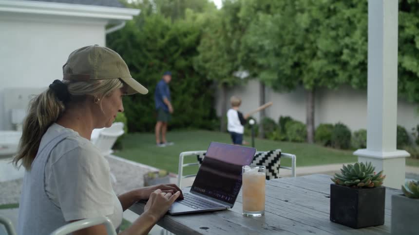 A woman sits outside her home and takes a sip of a drink while she uses her laptop and smiles as she watches her husband and son play baseball