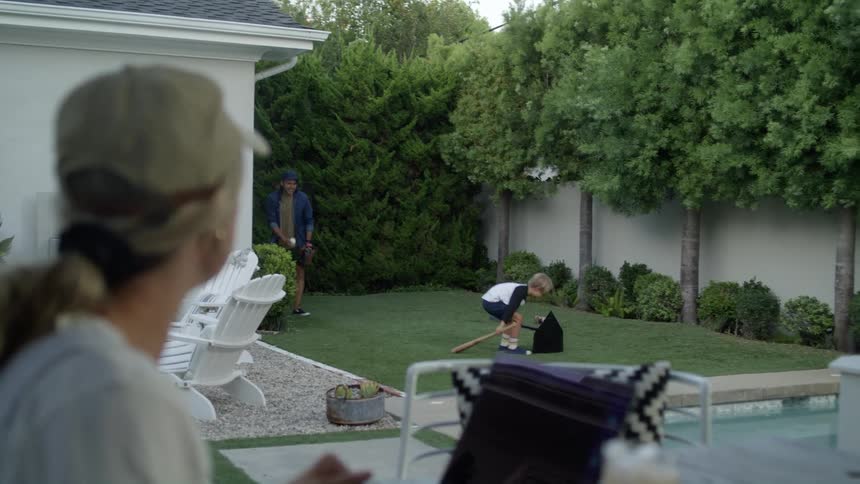 Mother types on laptop at outdoor table while watching father and son play t ball