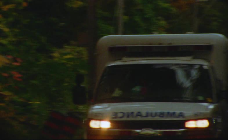 An ambulance with its lights flashing drives past a forest and a car