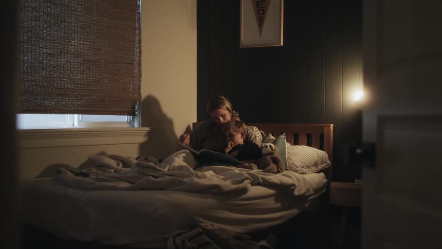 A woman sits in bed with her young son and reads a book to him