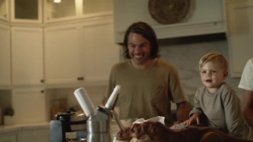 A little boy is standing in the kitchen with his family for breakfast and a bowl of waffles with a big pile of whipped cream on top when he grabs a handful of whipped cream his parents are laughing