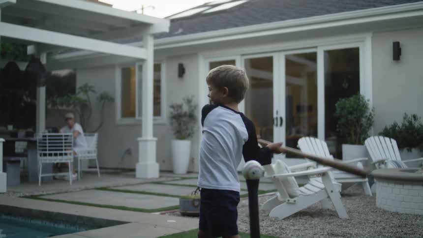 A man is outside in the yard teaching his young son how to hit a baseball as his mother watches from the patio
