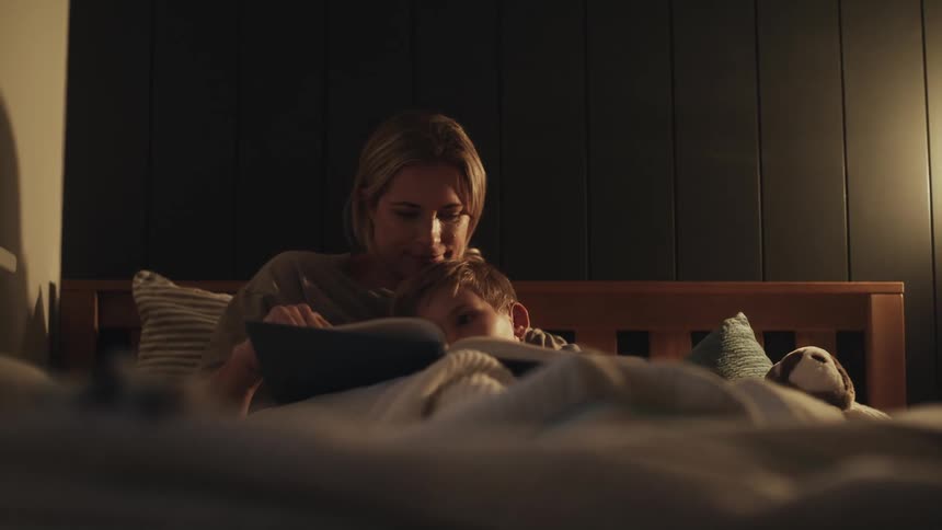 Mother sits in bed cuddling with son as she reads him a story