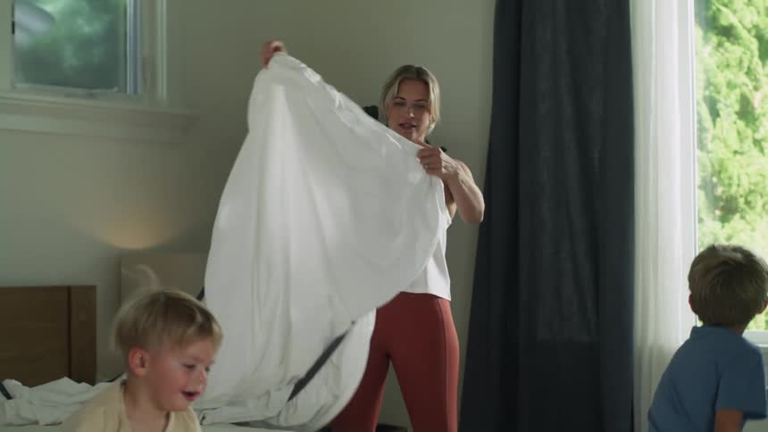 Mother and father are tying to put the sheet on the bed and one of their boys throws a pillow at his mother and she laughs