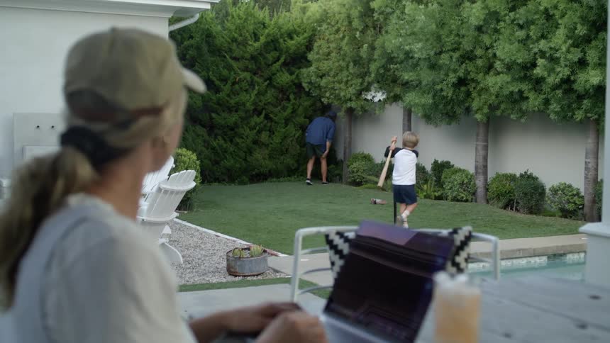 A woman works on her laptop while watching her husband and son play baseball in their backyard